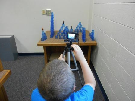A kid aims a camera on a tripod at stacks of cups with small figures on top.